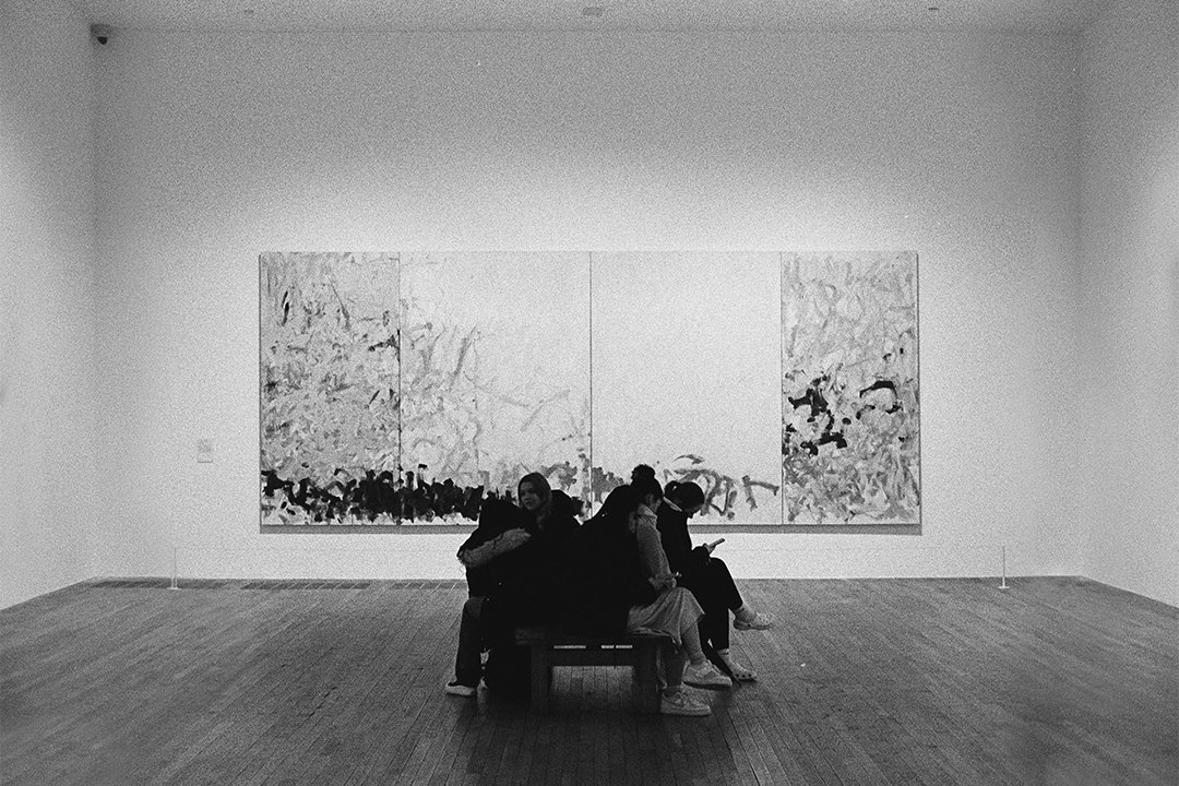 A black and white photo of a group of students sitting in a bunch, looking at their phones in front of a large gestural abstract painting.