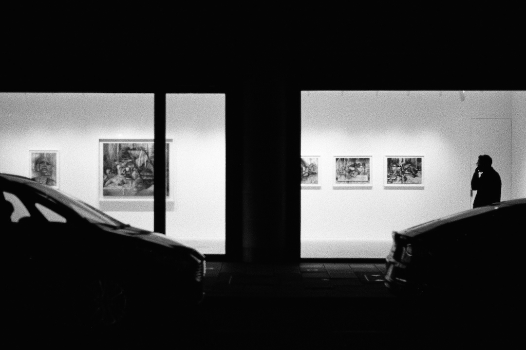A black and white photo of a modern art gallery at night. It's lights shine bright and put passers-by and cars in strong silhouette.