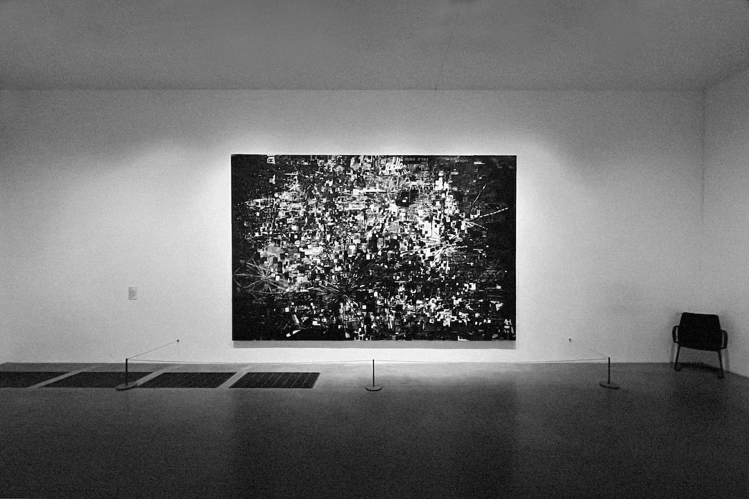 A black and white photo of a large abstract painting hanging in an art gallery with an empty invigilator's chair to the side.