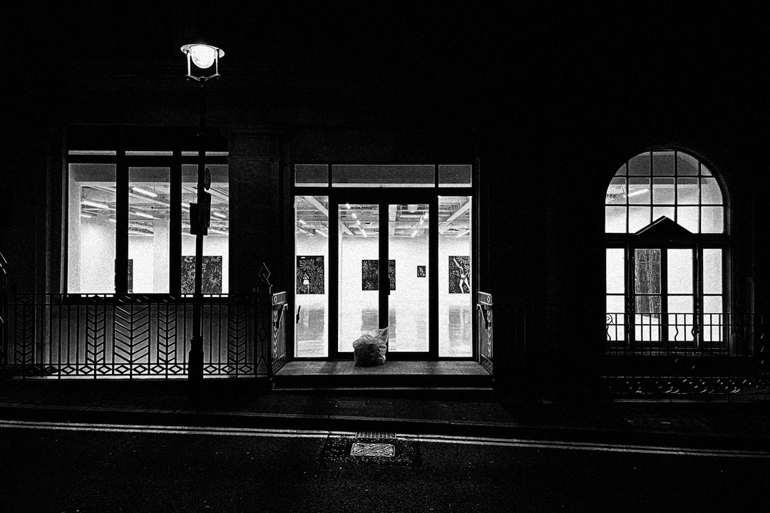 A strong contrast black and white photo of an art gallery at night. The bright lights illuminate the road and a refuse sack.