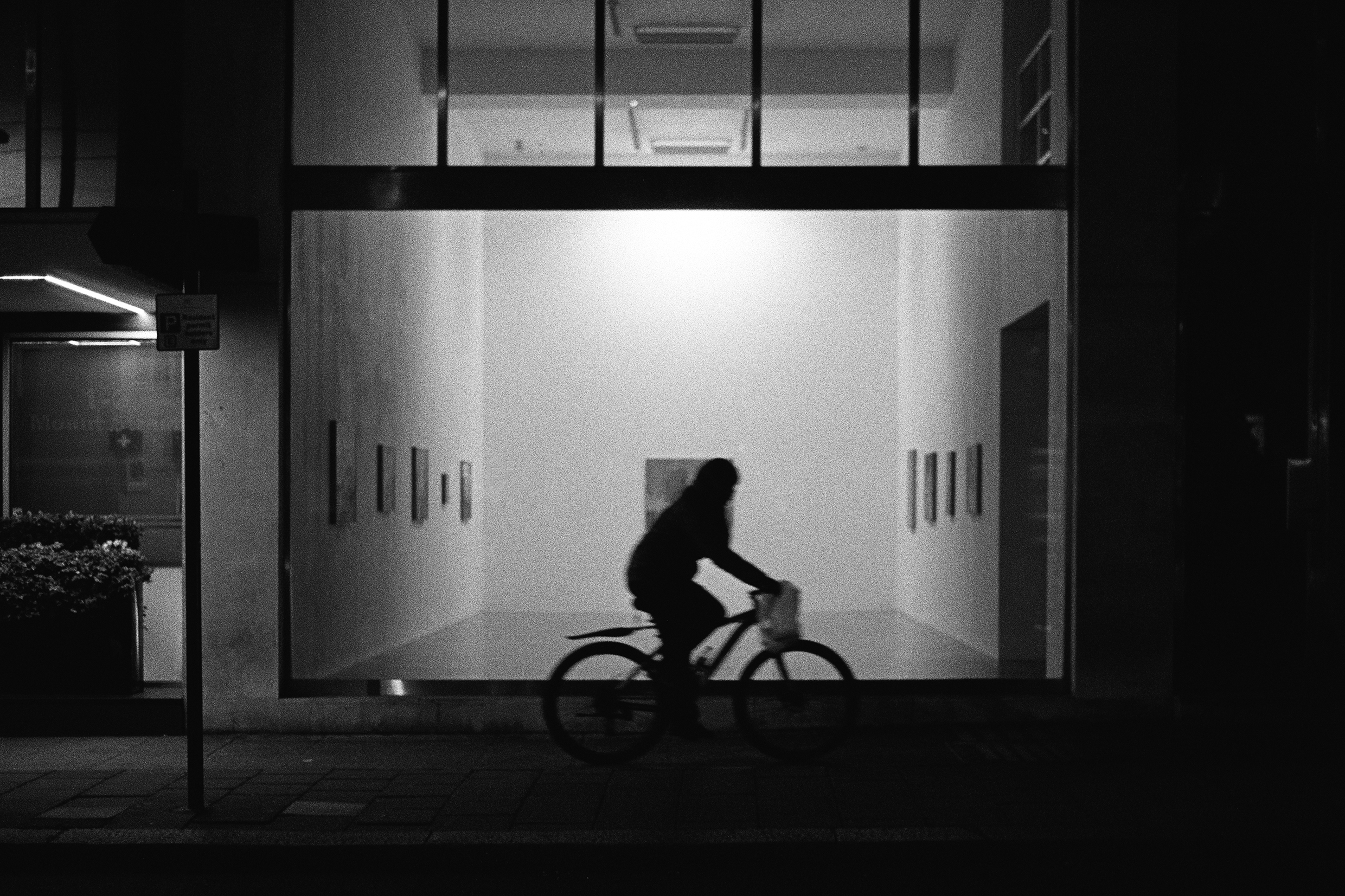 A black and white photo of a cyclist passing a modern art gallery at night. The bright lights of the gallery illuminate the scene and cast the cyclist into shadow.